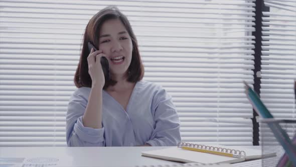 Young smiling asian woman working on laptop while sitting using phone for work in her home. alt