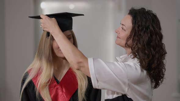 Smiling Mother Putting Graduation Cap Tassel Right to Left on Cap of Happy Daughter Standing Indoors alt