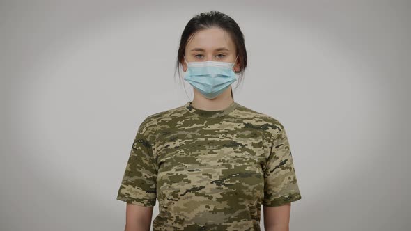 Front View Portrait of Serious Young Military Woman in Coronavirus Face Mask Posing at Grey alt