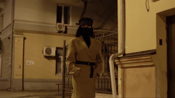 Girl in Black Virus Mask in a Beige Coat Walks Along Building in the Night City alt
