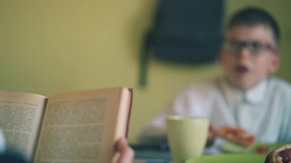 Schoolboy Reads Books Opposite Friend Eating Pizza at Table alt
