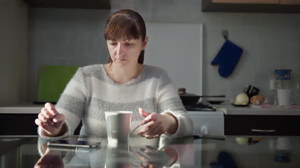 Sad Woman Sitting in the Kitchen at Home with a Cup of Tea and Uses the Phone Play a Game