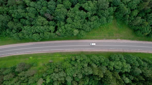 Road trip through a green deciduous forest. Aerial top view of cars driving along a scenic forest. alt