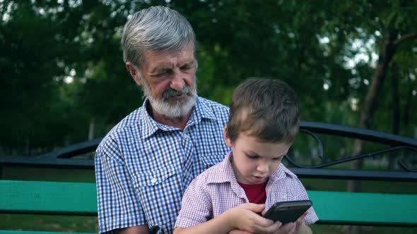 Senior Grandfather and Grandson Are Sitting on a Bench in the Park and Playing on a Smartphone alt