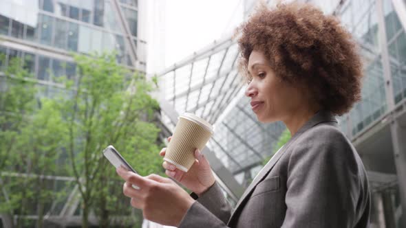 Afro American businesswoman in city using smart phone with takeaway coffee