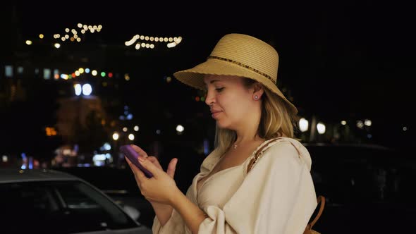 Handheld Shot of a Woman Using a Smartphone While Standing in the Middle of a Night City Street alt