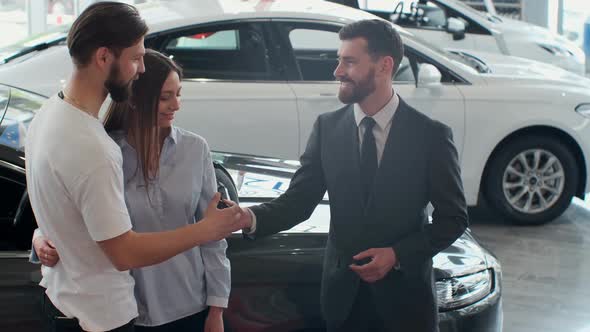 Successful Deal Between Car Salesman and Young Couple in Car Dealership alt