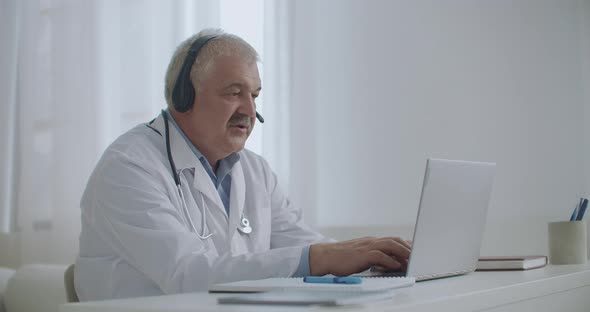 Male Doctor Is Talking with Patient Online By Video Call on Laptop, Listening By Headphones and alt