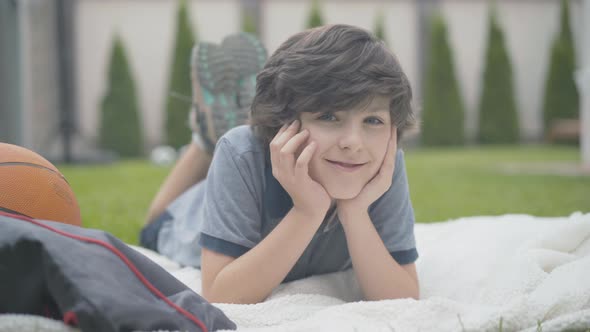 Portrait of Happy Kid Lying on Blanket Outdoors and Smiling at Camera. Joyful Relaxed Caucasian Boy alt