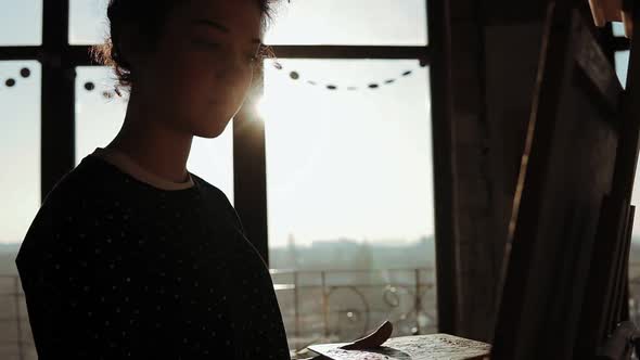 Shaded By the Sun Behind Young Talented Female Artist Drawing on Easel in an Art Studio alt