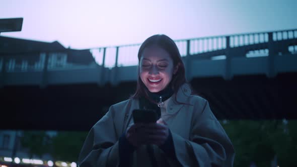 Young woman surrounded with evening city lights she is looking at her phone and laughs alt