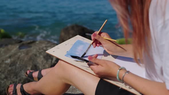 Female Student of School of Art is Painting Marine Landscape Sitting on Coast alt