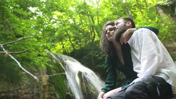 Young Couple - Man with Beard and Woman with Curly Hair Is Hugging and Kissing By Sitting on Stone alt