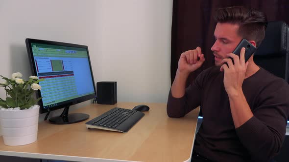 A Young, Handsome Man Sits in Front of a Computer Screen and Discusses on a Smartphone alt