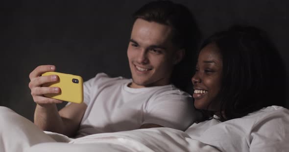 Happy Multi-ethnic Couple Watching Video Together on the Yellow Smartphone Lying in Bed at Night alt