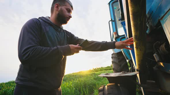 Man in Field with Phone in His Hands Stands By His Tractor and Knocks on Wheel Checking Condition of alt
