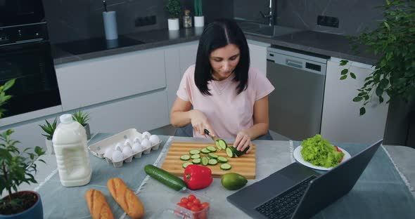Brunette in Modern Cuisine and Cutting Cucumber for Salad Simultaneously Choosing Program on Laptop alt