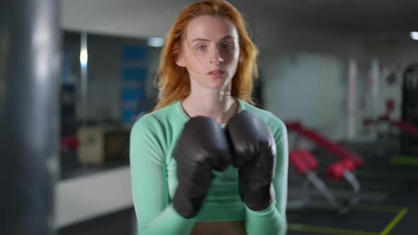 Perspiring Satisfied Redhead Sportswoman Hitting Boxing Gloves Looking at Camera with Blurred Woman alt