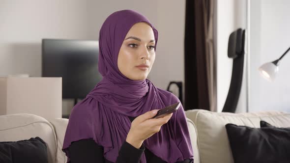A Muslim Woman Watches TV That's Off Camera and Switches Through Channels with a Remote Control alt