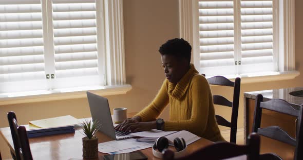 African american woman using laptop and reading documents while working from home alt