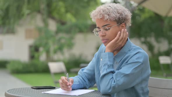 Young African Woman Thinking While Writing on Paper in Outdoor Cafe alt