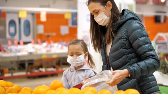 Young woman with child girl in medical masks buys a oranges at supermarket