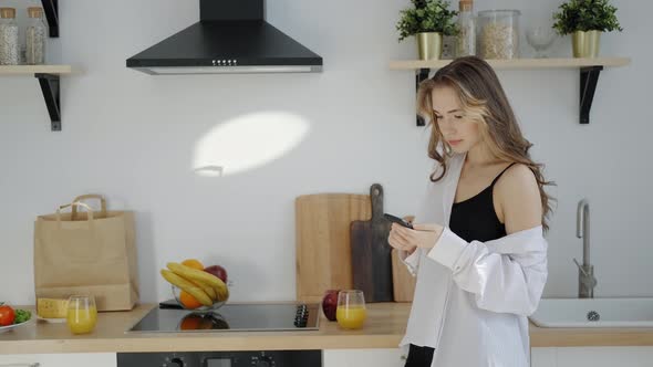 Young Woman in White Men's Shirt with Smartphone in Morning Kitchen