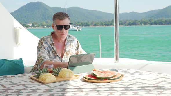 4K Caucasian businessman working on digital tablet while catamaran boat sailing on summer vacation. alt