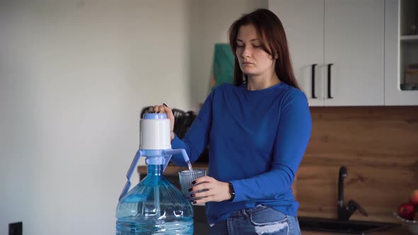 Woman Presses Pumps Water From a Large Bottle Into a Blue Glass at Home Kitchen