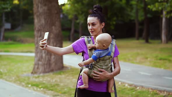 Young woman taking selfie while carrying baby in sling alt