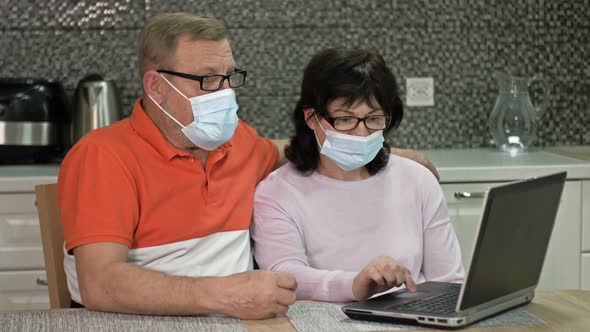 Couple Senior in Protective Masks Using Laptop at Home for Online Shopping alt