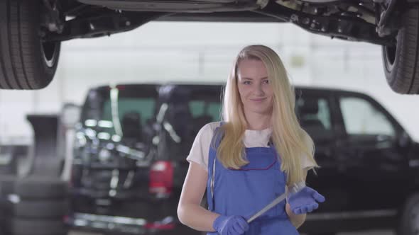 Portrait of Young Female Auto Mechanic in Workrobe Standing Under Car in Repair Shop, Looking at alt