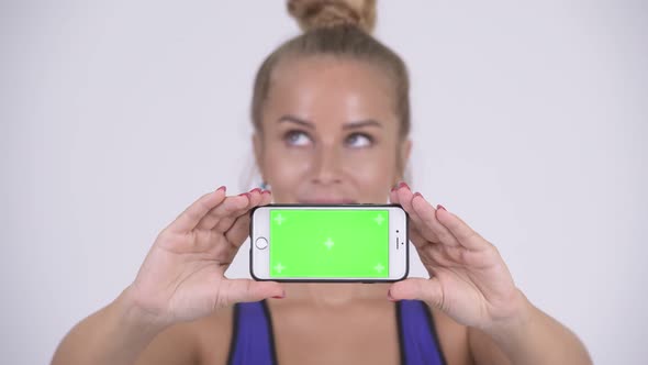Face of Happy Young Blonde Woman Thinking While Showing Phone alt