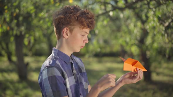 Side View Handsome Teenage Boy with Red Hair Launching Paper Plane Outdoors Looking Away alt