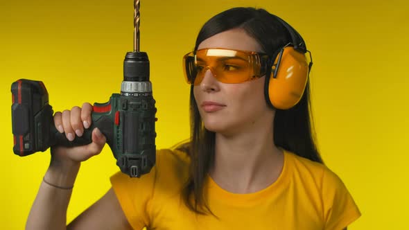 A Young Brunette Woman in Protective Headphones and Glasses is Holding a Drill alt