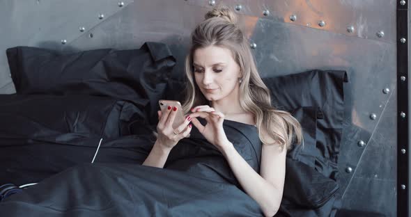 Gorgeous Blondie Checking Her Mobile Phone Sitting in Bed Under Black Blanket