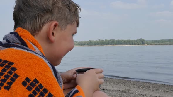 a Teenage Guy Sits on the Bank of the River Wrapped in a Towel and Drinks Hot Tea From a Metal Mug