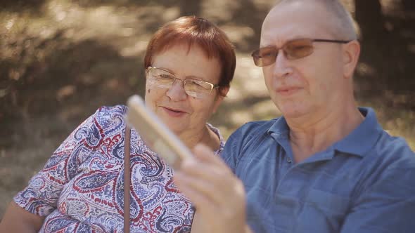 Elderly Couple on a Park Bench Taking Selfies on Smartphone alt