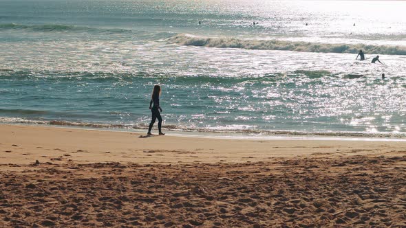 Female surfer walking down the Atlantic Ocean beach at sunset. alt