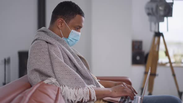 Side View of Concentrated African American Man in Covid Face Mask Coughing Typing on Laptop Keyboard alt
