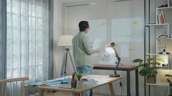 Back View Of Asian Man Designer Compare The Bag Design Sketch Paper In Hand To The Paper On The Wall alt