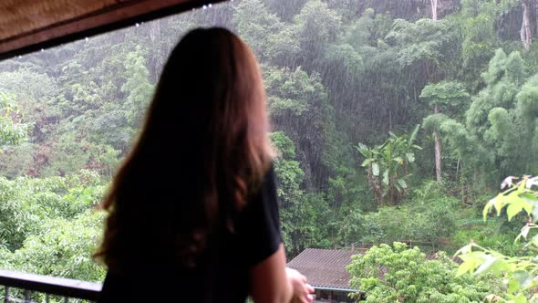 a woman standing on balcony and looking at the rain over the tree