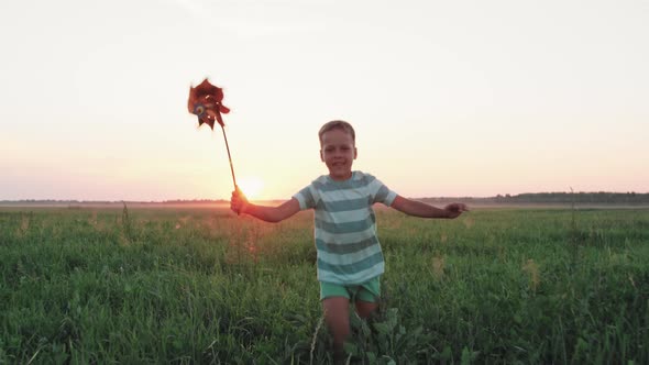 Child Boy Running with a Toy Windmill Across a Field in Summer at Sunset alt