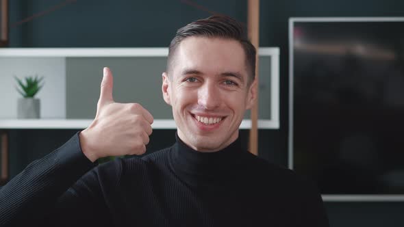 Positive Young Man Smiling to Camera and Showing Thumb Up Sign at Home Office Background