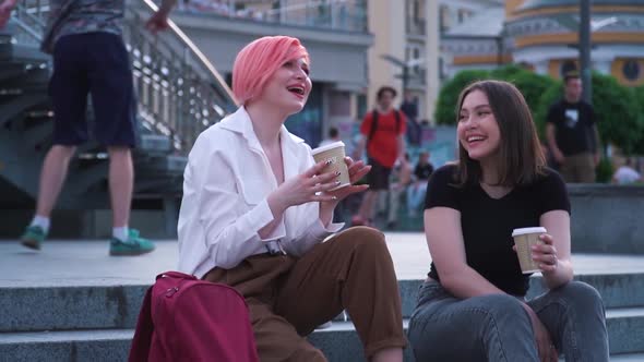Two Young Women Talking and Laughing Sitting on the Granite Stairs and Drinking Coffee alt