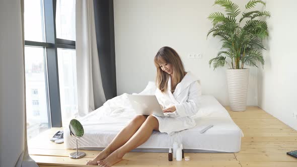 Young Chinese Woman Working at a Laptop Online She Is Sitting on the Bed in a White Coat at a Laptop alt