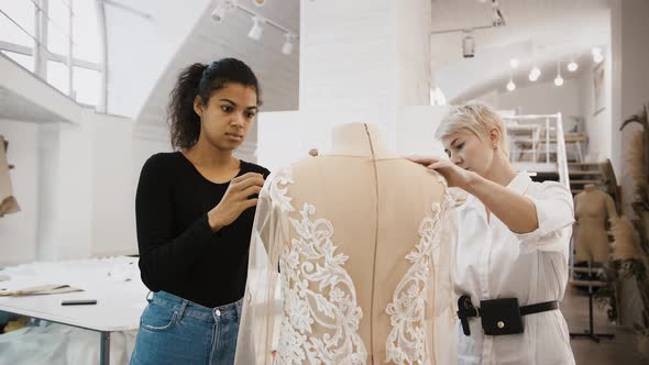 Females Tailors are Working on Wedding Dress Hanging on Mannequin Measuring and Pinning It Up alt