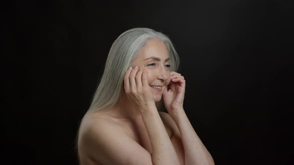 Beautiful Grey Haired Senior Lady with Bare Shoulders Doing Facial Self Massage Black Background alt