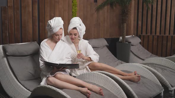 Two Young Relaxed Women Looking at Menu in Spa Salon Choosing Procedures