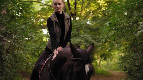 Atractive Woman Riding Beautiful Brown Horse in Park During Sunny Day and Stroking the Horse's Head alt